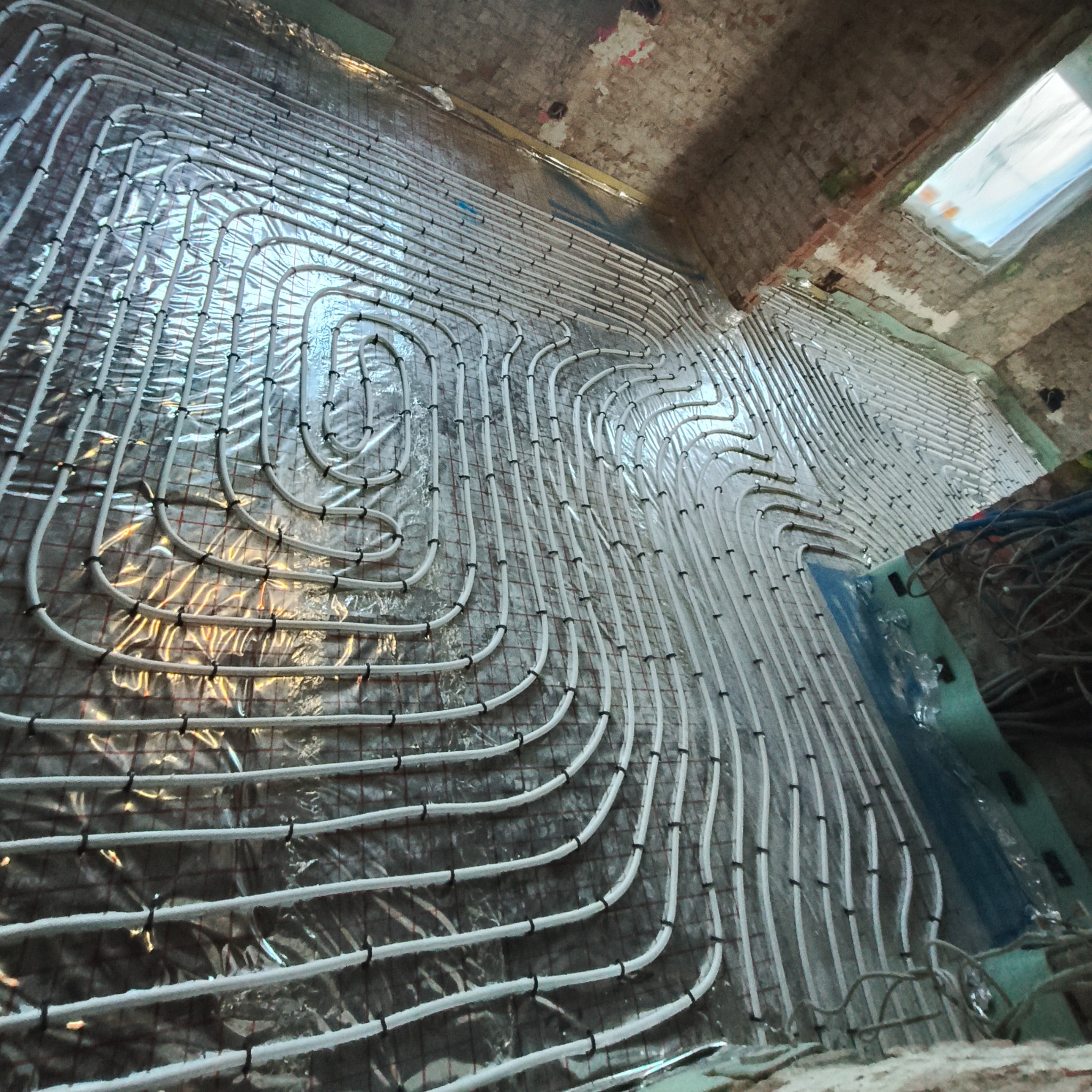 Białe rury ogrzewania podłogowego ułożone spiralnie na srebrnej folii w Krowiarkach, przymocowane czarnymi klipsami do siatki. Widok z góry, surowy stan.