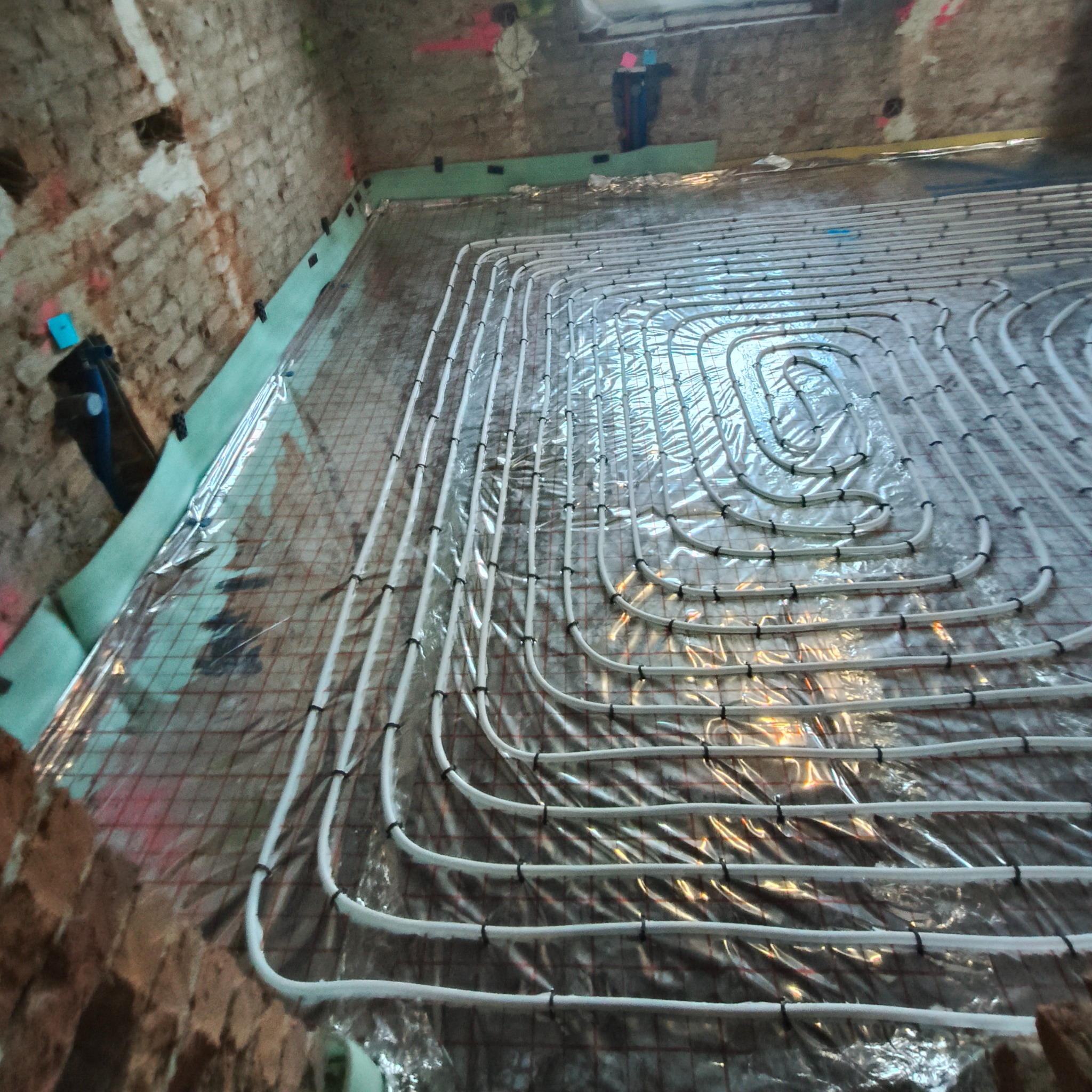 Białe rury ogrzewania podłogowego ułożone na siatce w surowym wnętrzu z ceglanymi ścianami. Instalacja przed wylewką, widok z góry.