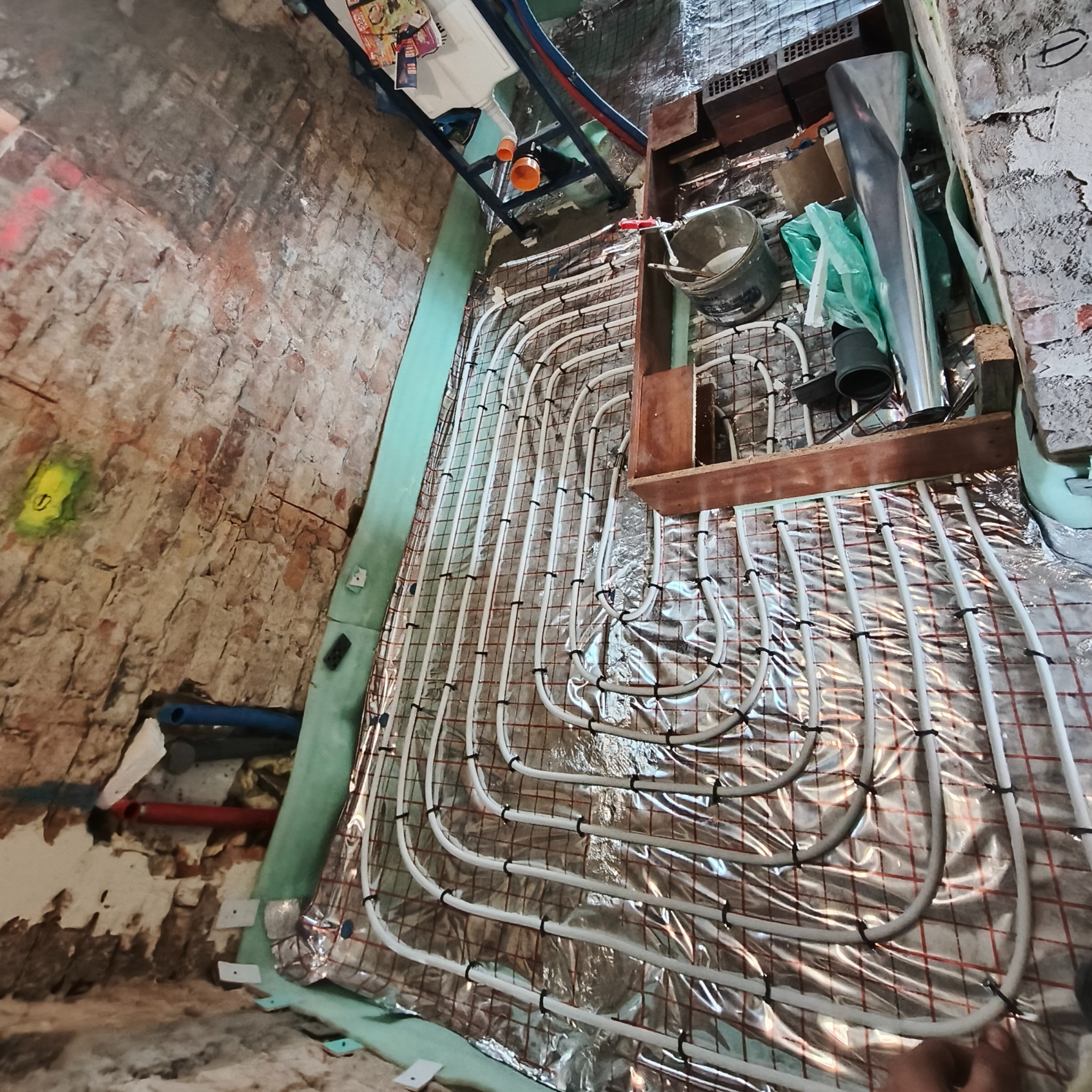 Układanie rur ogrzewania podłogowego na folii izolacyjnej z siatką, widok z góry, surowa ściana z cegły w tle, etap prac przed wylewką.