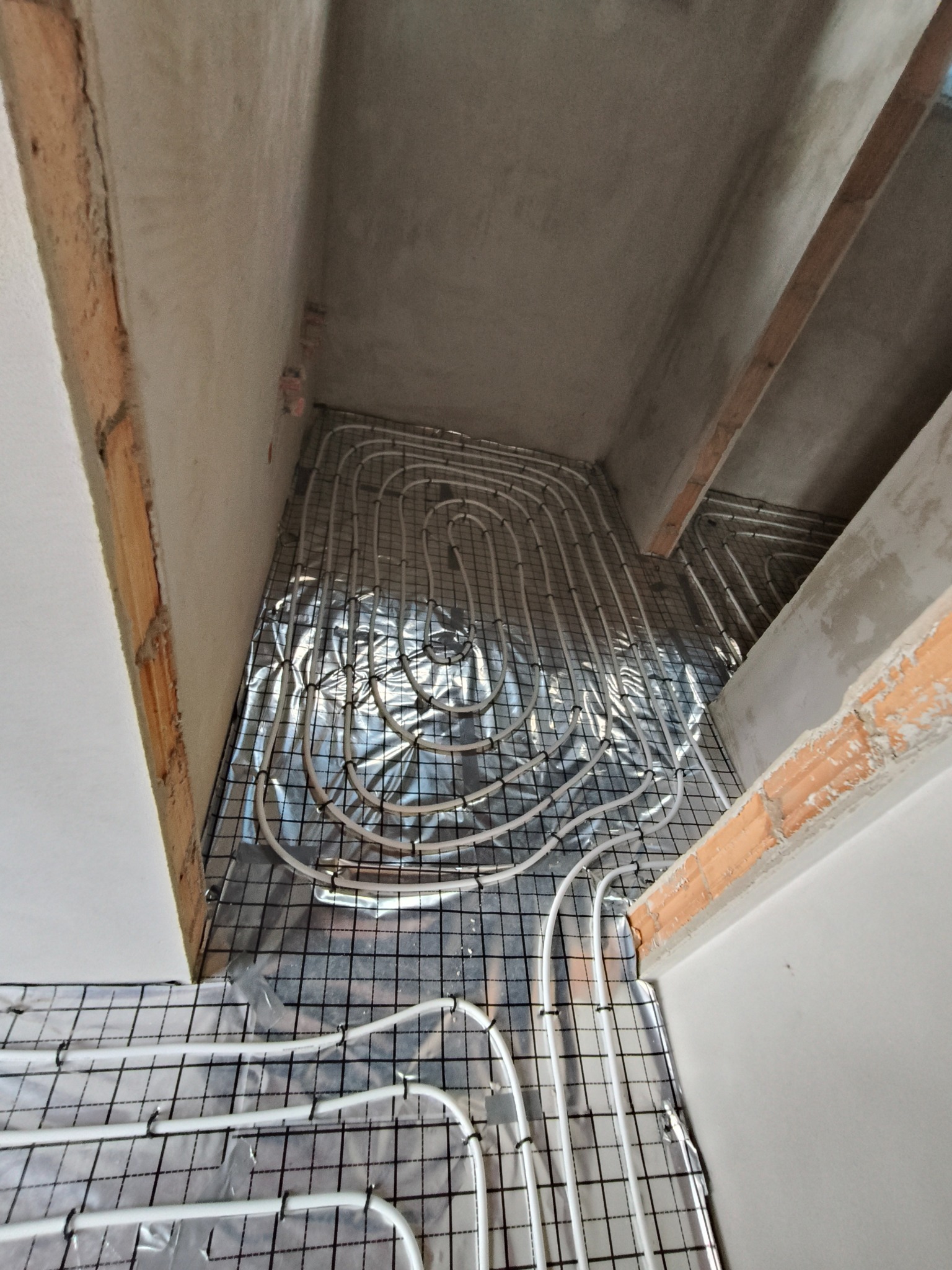 Białe rury ogrzewania podłogowego ułożone na folii odblaskowej i siatce w surowym wnętrzu. Widoczne fragmenty ścian z cegły i betonu, instalacja w trakcie realizacji.