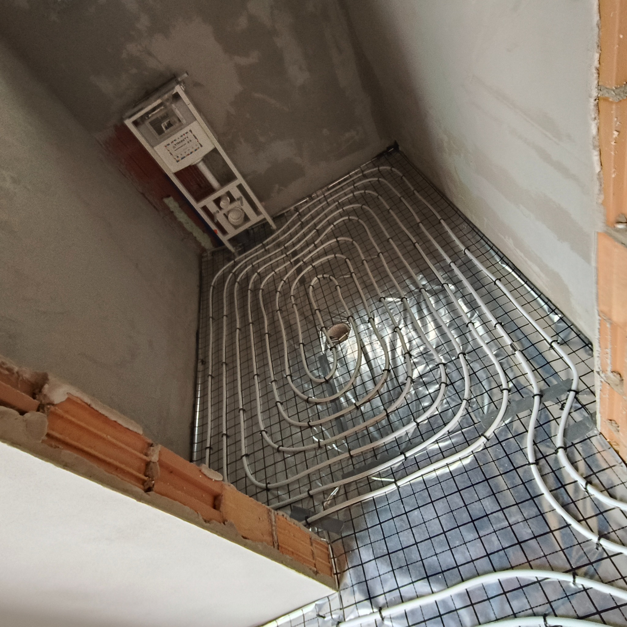 Białe rury ogrzewania podłogowego ułożone na czarnej siatce, widoczne fragmenty surowych ścian i instalacji sanitarnych w surowym wnętrzu.