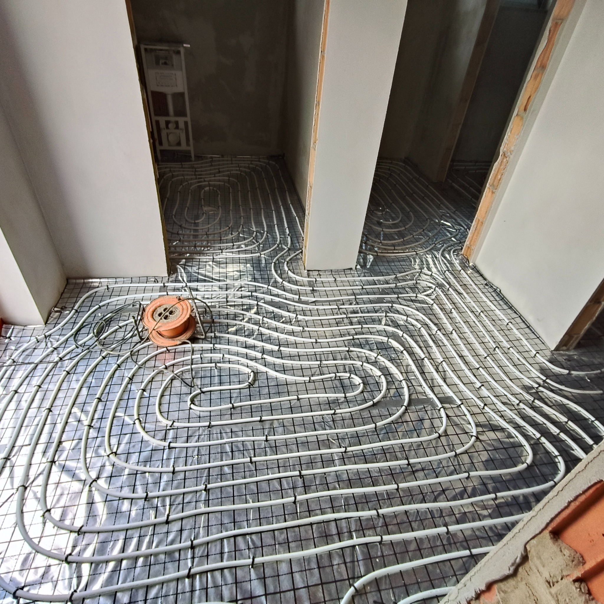 Białe rury ogrzewania podłogowego ułożone na metalowej siatce, widoczne w surowym wnętrzu budynku w trakcie instalacji. Pomarańczowa rolka z rurami obok.