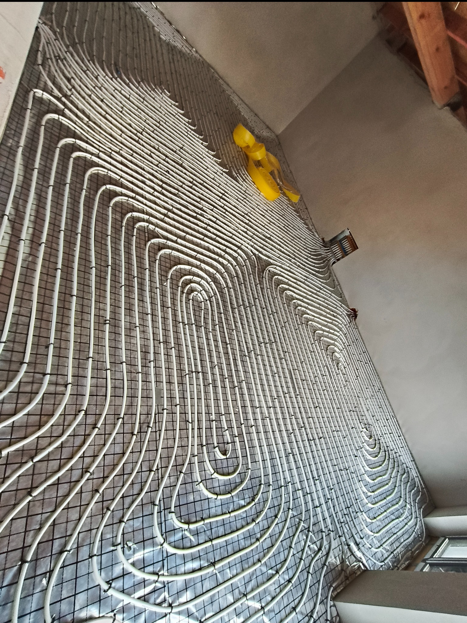 Białe rury ogrzewania podłogowego ułożone na czarnej siatce, z żółtą taśmą zabezpieczającą, widoczne pod kątem w surowym wnętrzu.