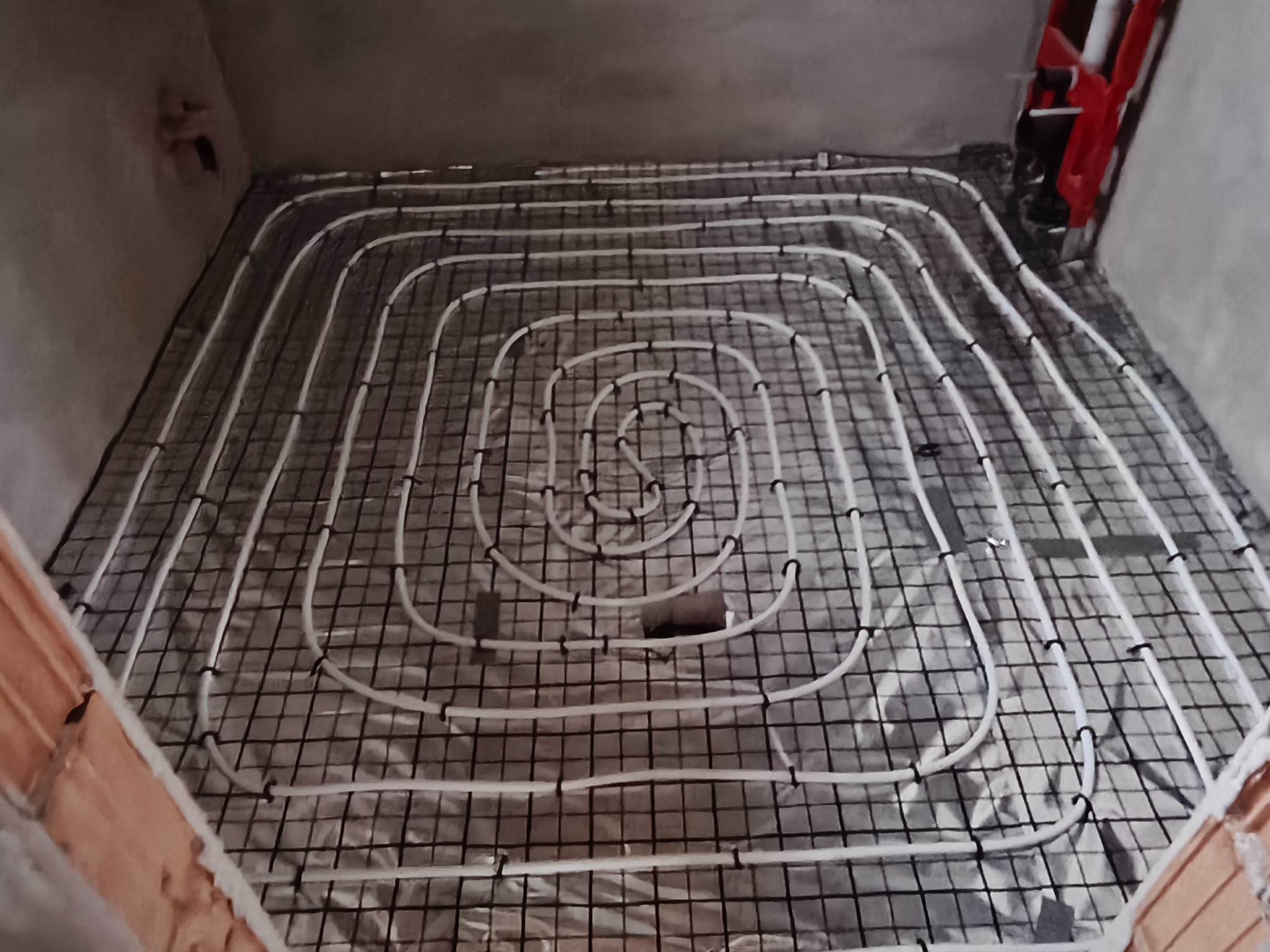 Ułożone białe rury ogrzewania podłogowego na siatce w surowym pomieszczeniu, widoczne fragmenty ścian. Realizacja instalacji grzewczej.
