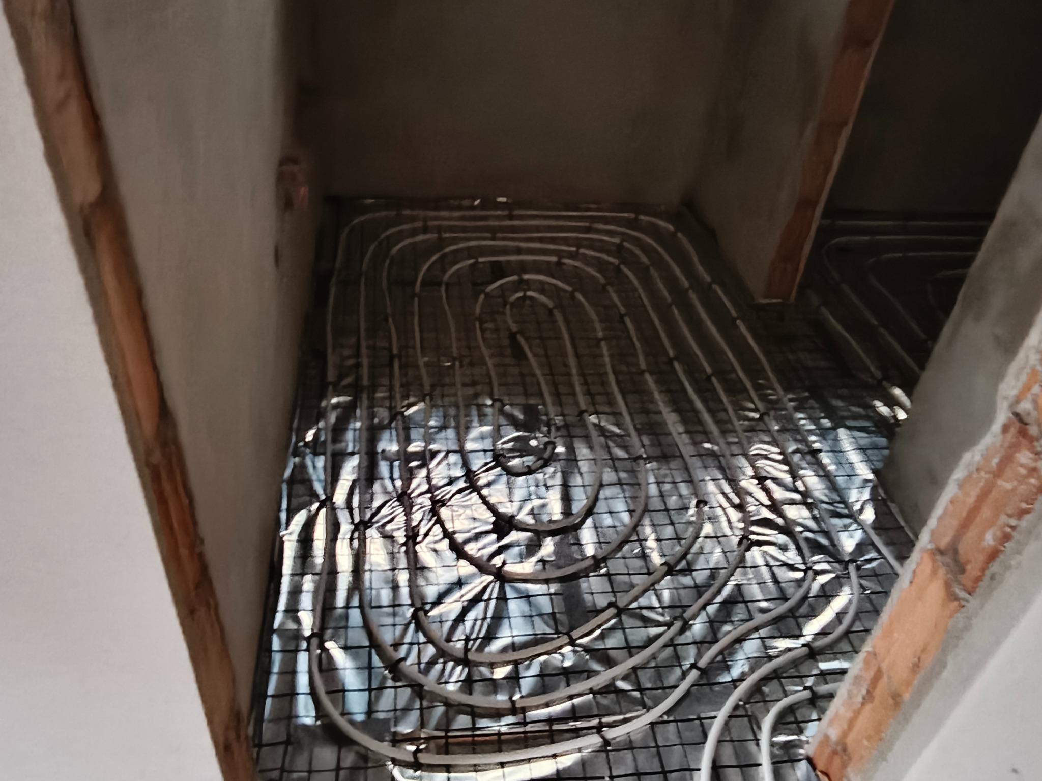 Białe rury ogrzewania podłogowego ułożone na srebrnej folii na czarnej siatce, widok z góry, surowy stan wykończenia ścian i podłogi.