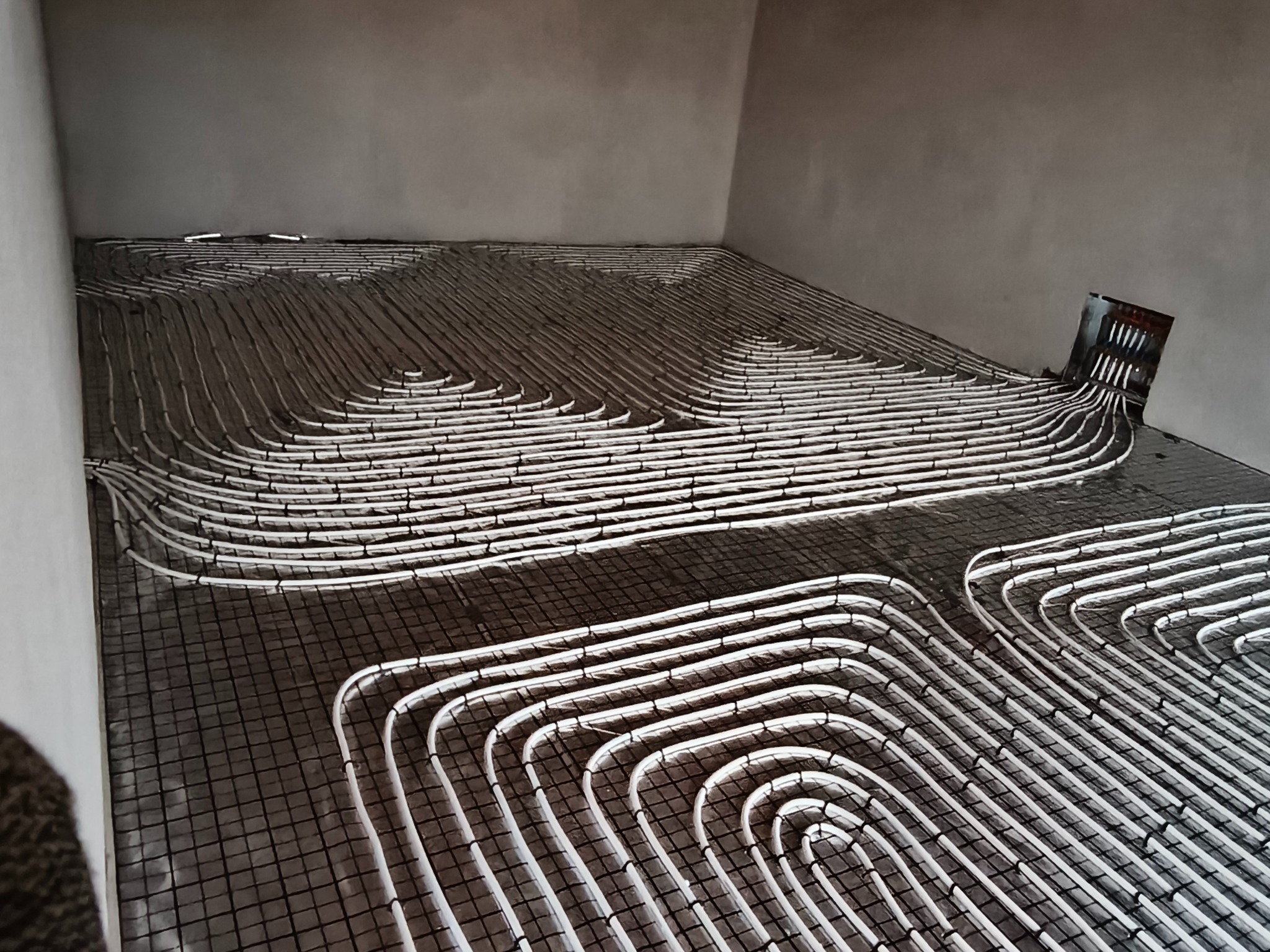 Białe rury ogrzewania podłogowego ułożone na czarnej siatce w surowym wnętrzu, gotowe do zalania wylewką. Widok z góry na rozłożoną instalację.