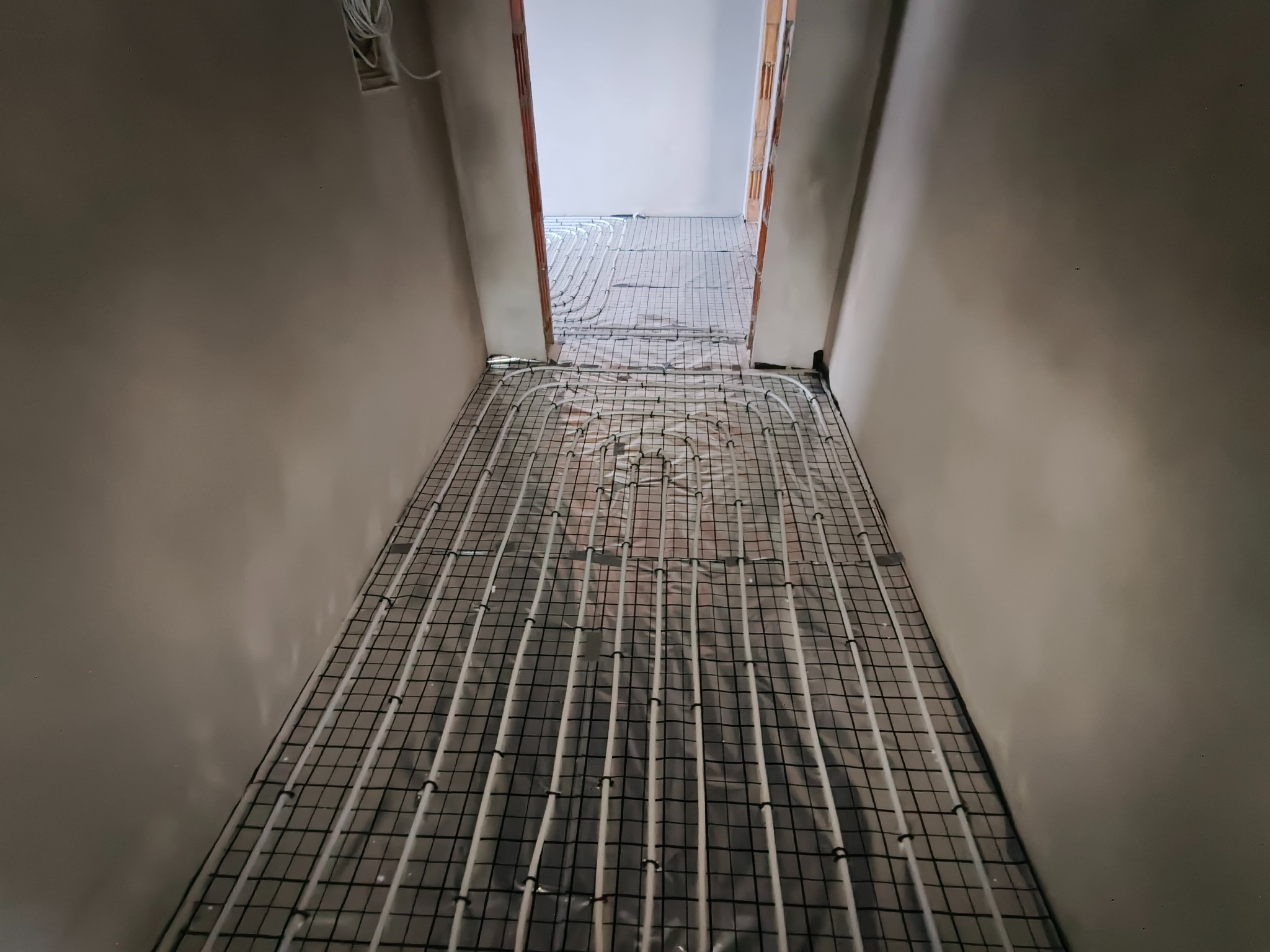 Białe rurki ogrzewania podłogowego ułożone na czarnej siatce w korytarzu, widok z góry. Instalacja przed wylaniem posadzki. Perspektywa zbieżna, neutralne kolory.