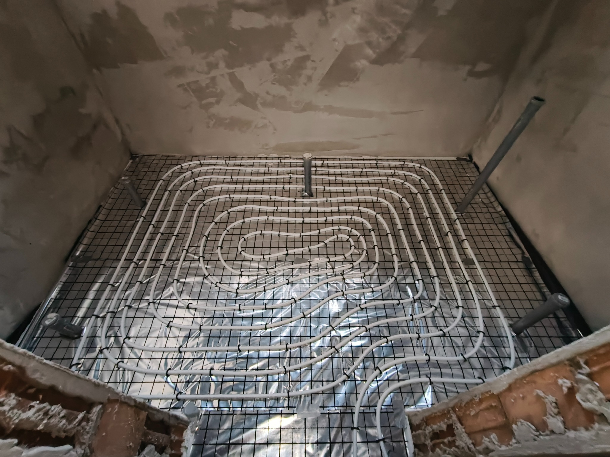 Białe rury ogrzewania podłogowego ułożone na folii i siatce w surowym wnętrzu, widok z góry na instalację przed wylewką. Elementy instalacji wystają ze ściany.