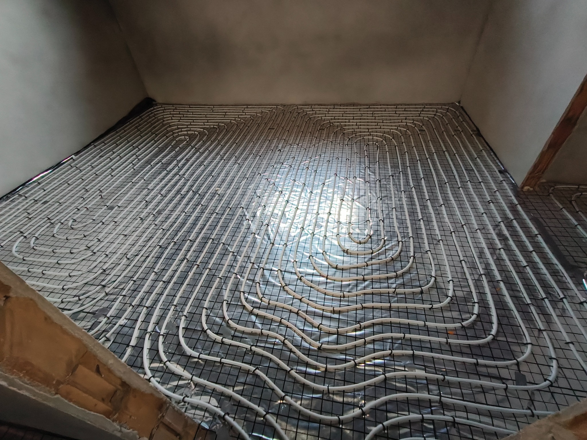 Białe rury ogrzewania podłogowego ułożone na czarnej siatce, widok z góry, przed wylaniem posadzki. Układ spiralny rur w jasnym pomieszczeniu w trakcie budowy.