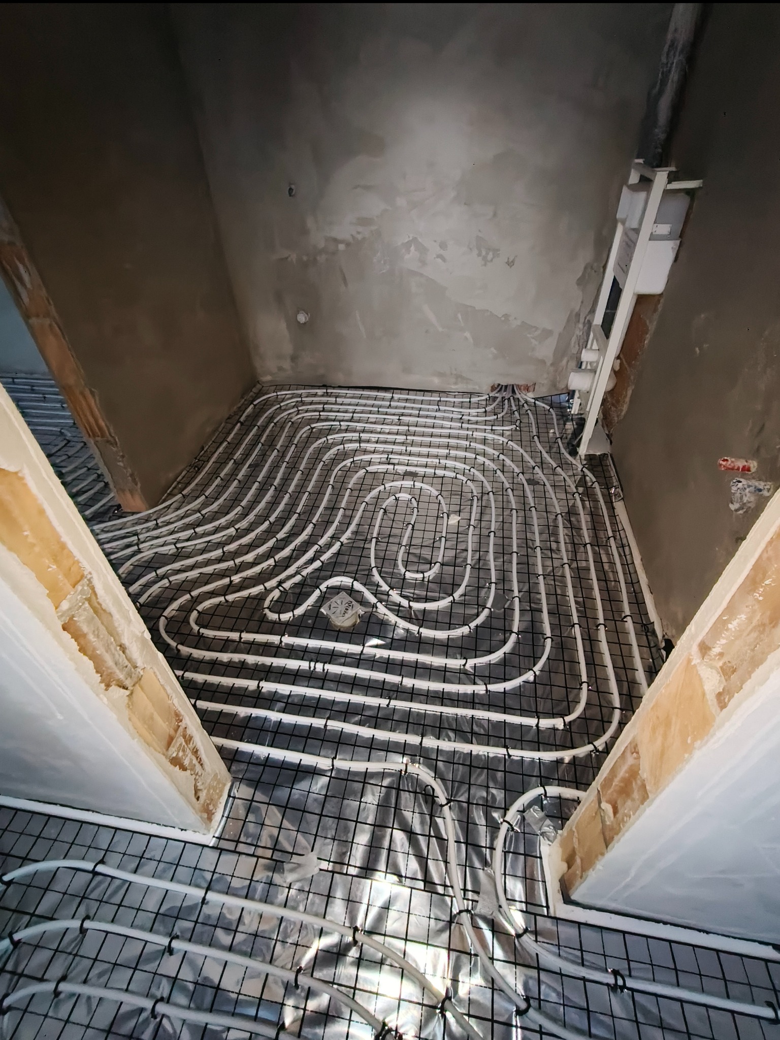 Białe rurki ogrzewania podłogowego ułożone na czarnej siatce w surowym wnętrzu łazienki, widok z góry na instalację przed wylaniem posadzki.