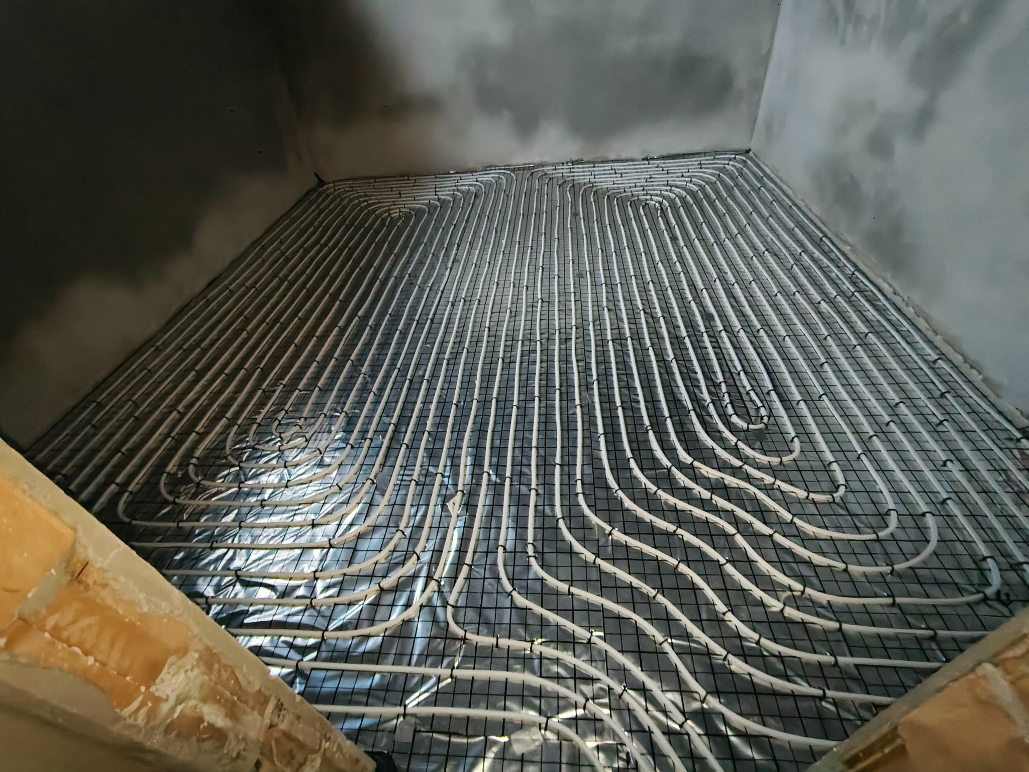 Białe rury ogrzewania podłogowego ułożone w pętle na czarnej siatce, widoczne fragmenty surowych ścian i podłoża w surowym stanie.