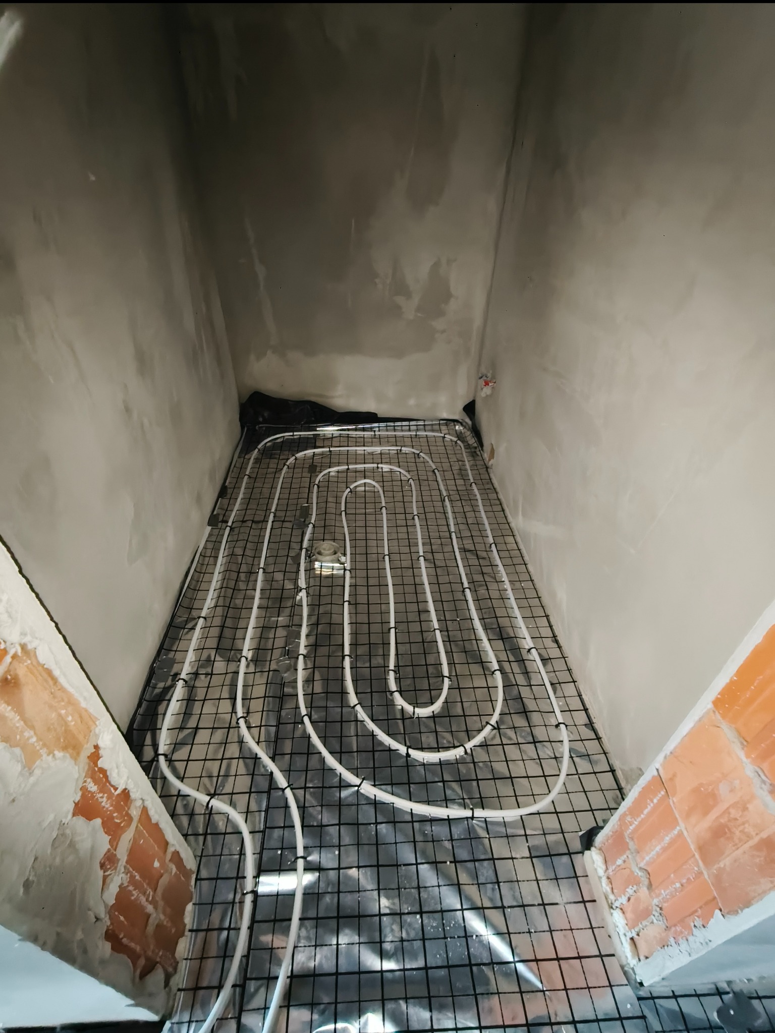 Białe rurki ogrzewania podłogowego ułożone na czarnej siatce w surowym wnętrzu. Widoczne ceglane fragmenty ścian i szare, nieotynkowane ściany.