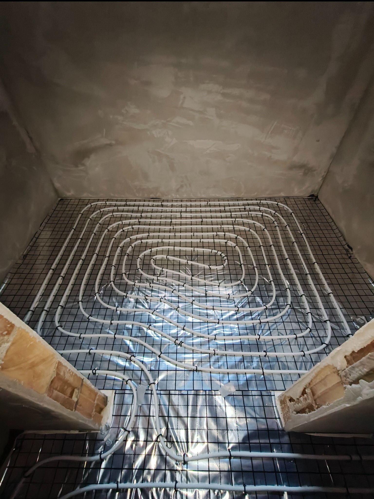 Białe rury ogrzewania podłogowego ułożone na czarnej siatce, widoczne z góry w surowym wnętrzu przed wylewką. Fragment ściany z cegły w dolnym kadrze.