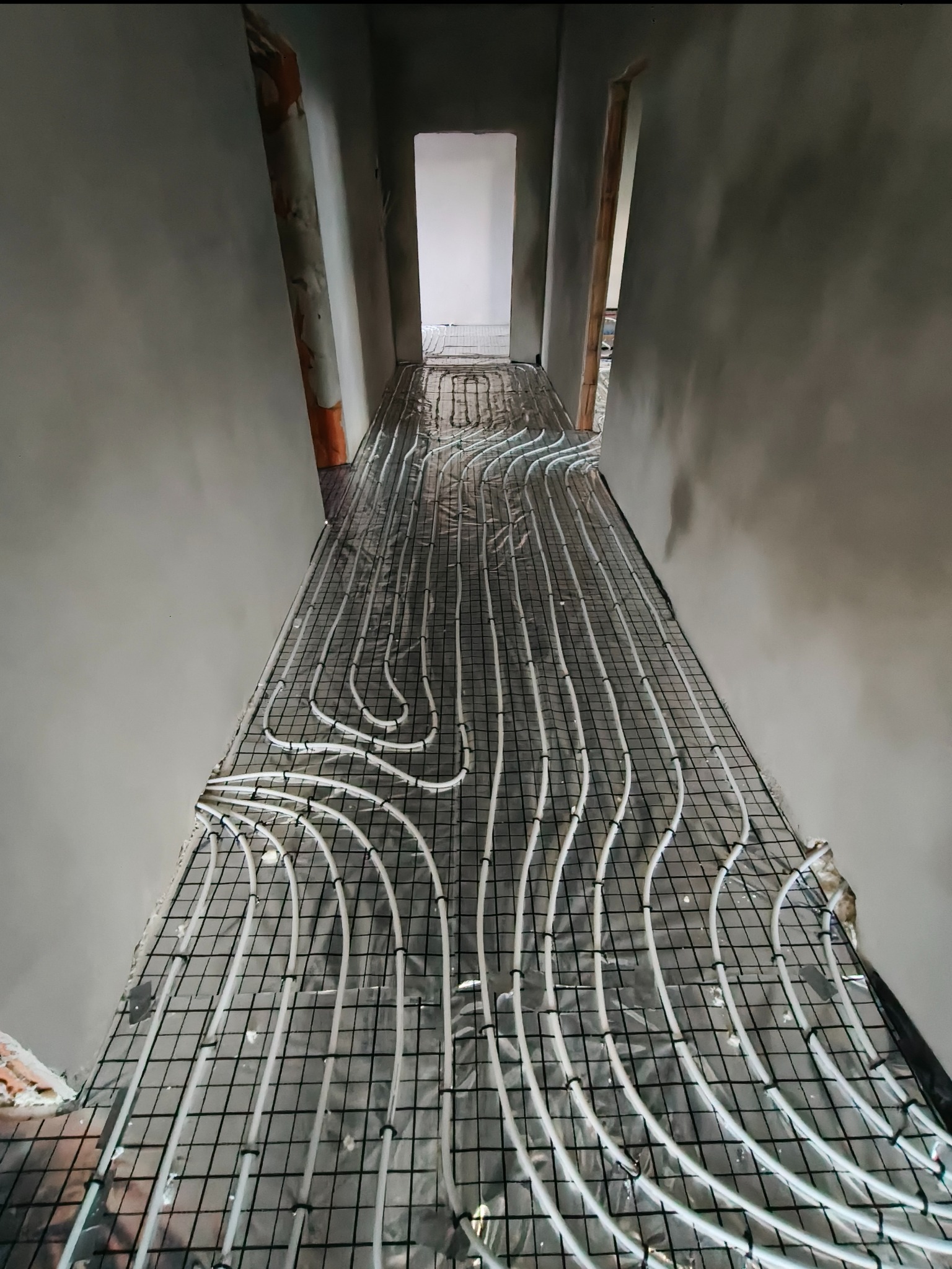 Układanie białych rur ogrzewania podłogowego na czarnej siatce w korytarzu z surowymi ścianami. Widok z góry na instalację przed wylaniem posadzki.