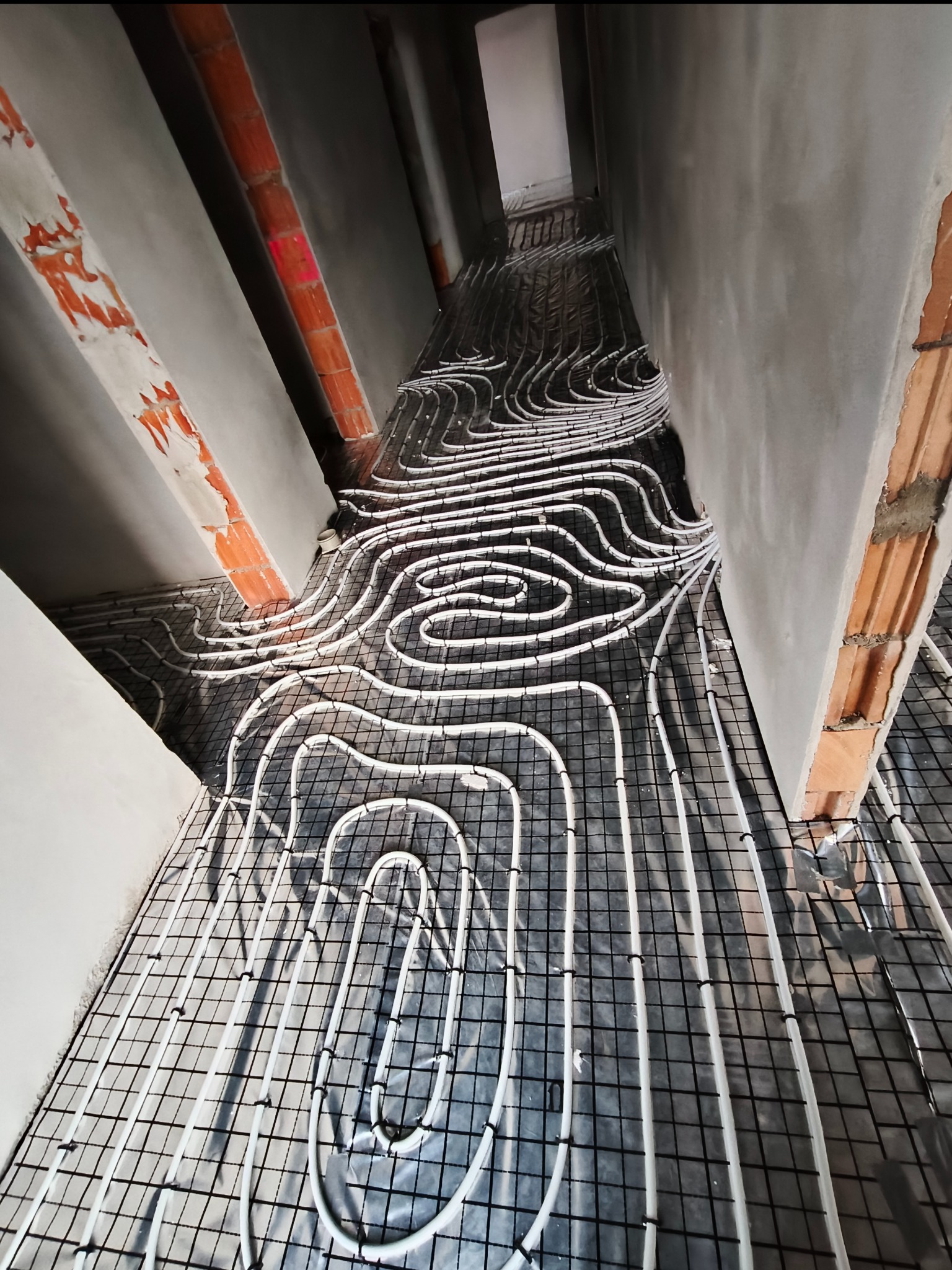 Białe rury ogrzewania podłogowego ułożone na czarnej siatce montażowej w surowym wnętrzu z ceglanymi elementami konstrukcyjnymi.