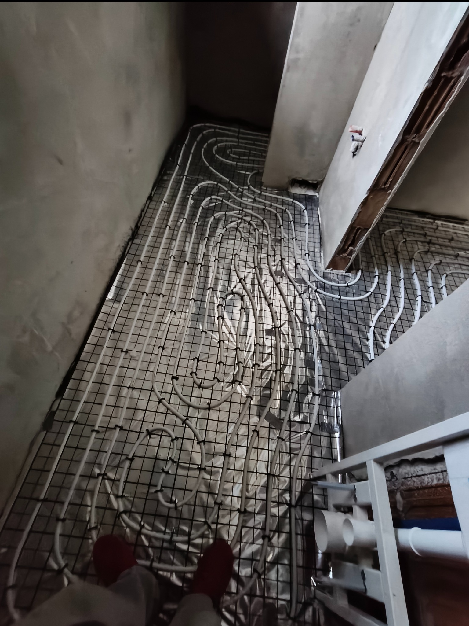 Białe rury ogrzewania podłogowego ułożone na siatce, widoczne fragmenty ścian w surowym stanie i instalacja sanitarna. Etap prac przed wylewką.