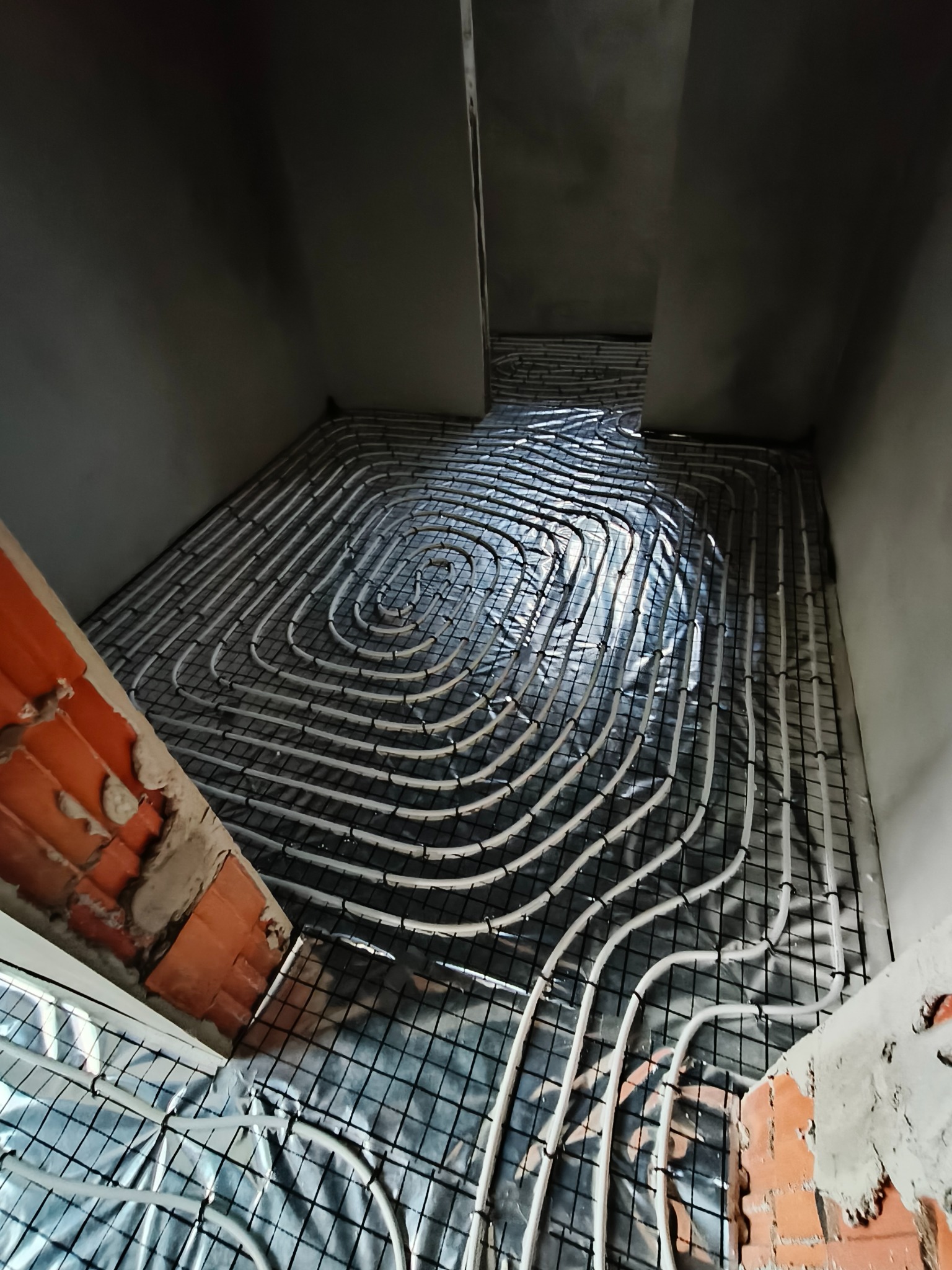 Układanie białych rur ogrzewania podłogowego na metalowej siatce, widok z góry w surowym wnętrzu z fragmentem ceglanej ściany. Etap instalacji przed wylewką.