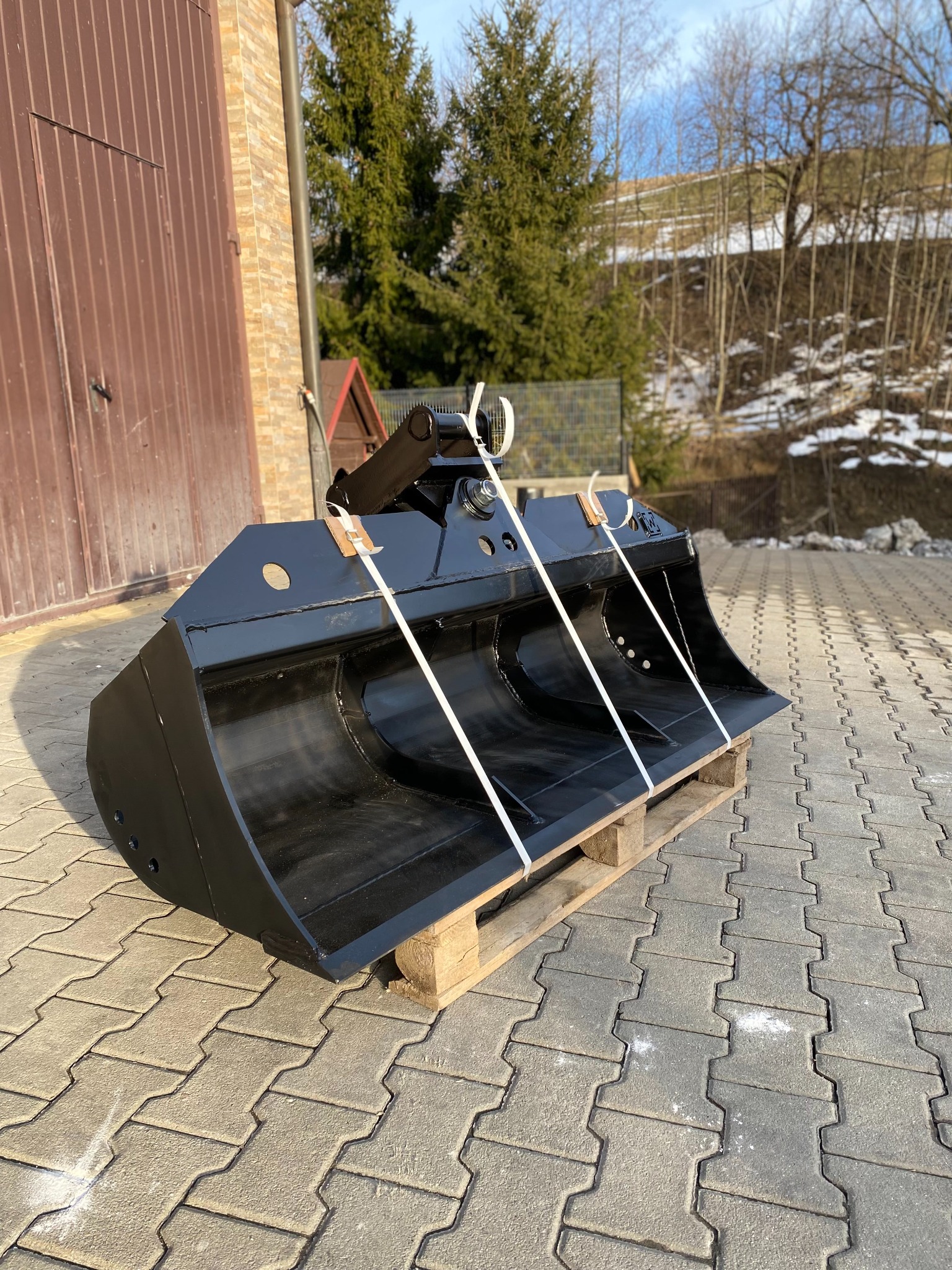 Nowa, czarna łyżka skarpowa hydrauliczna na drewnianej palecie, zabezpieczona białymi pasami transportowymi, na tle kostki brukowej, brązowego budynku gospodarczego i drzew z resztkami śniegu.