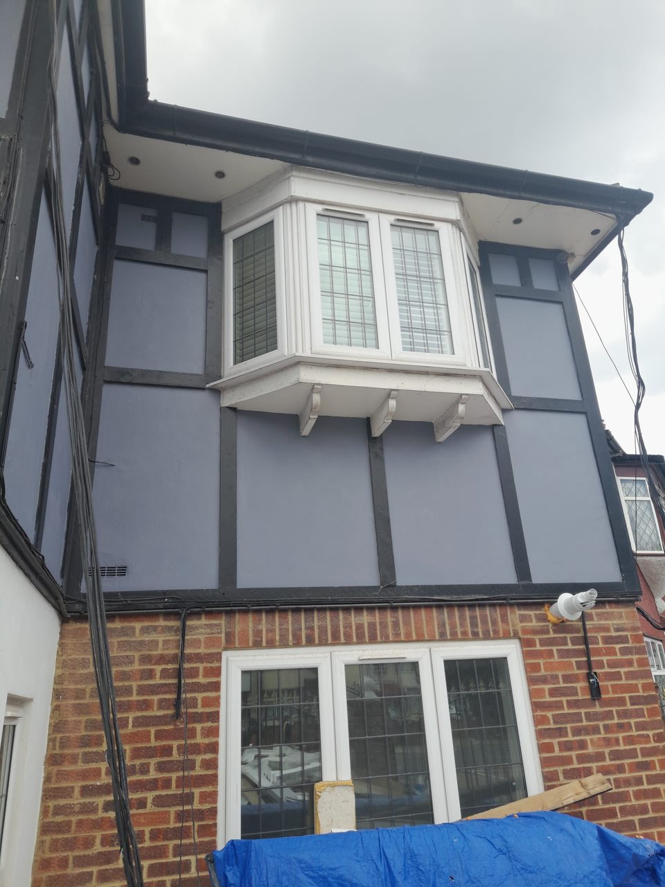 Odnowiona elewacja domu z szarymi panelami i czarnymi belkami, widoczny biały wykusz okienny i ceglana ściana z zabezpieczeniem niebieską folią.