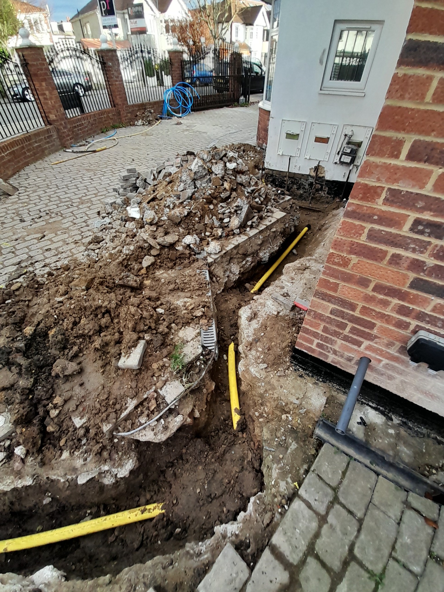 Wykop w ziemi z widocznymi żółtymi rurami instalacyjnymi biegnącymi wzdłuż fundamentów budynku, gruz i kostka brukowa na powierzchni.