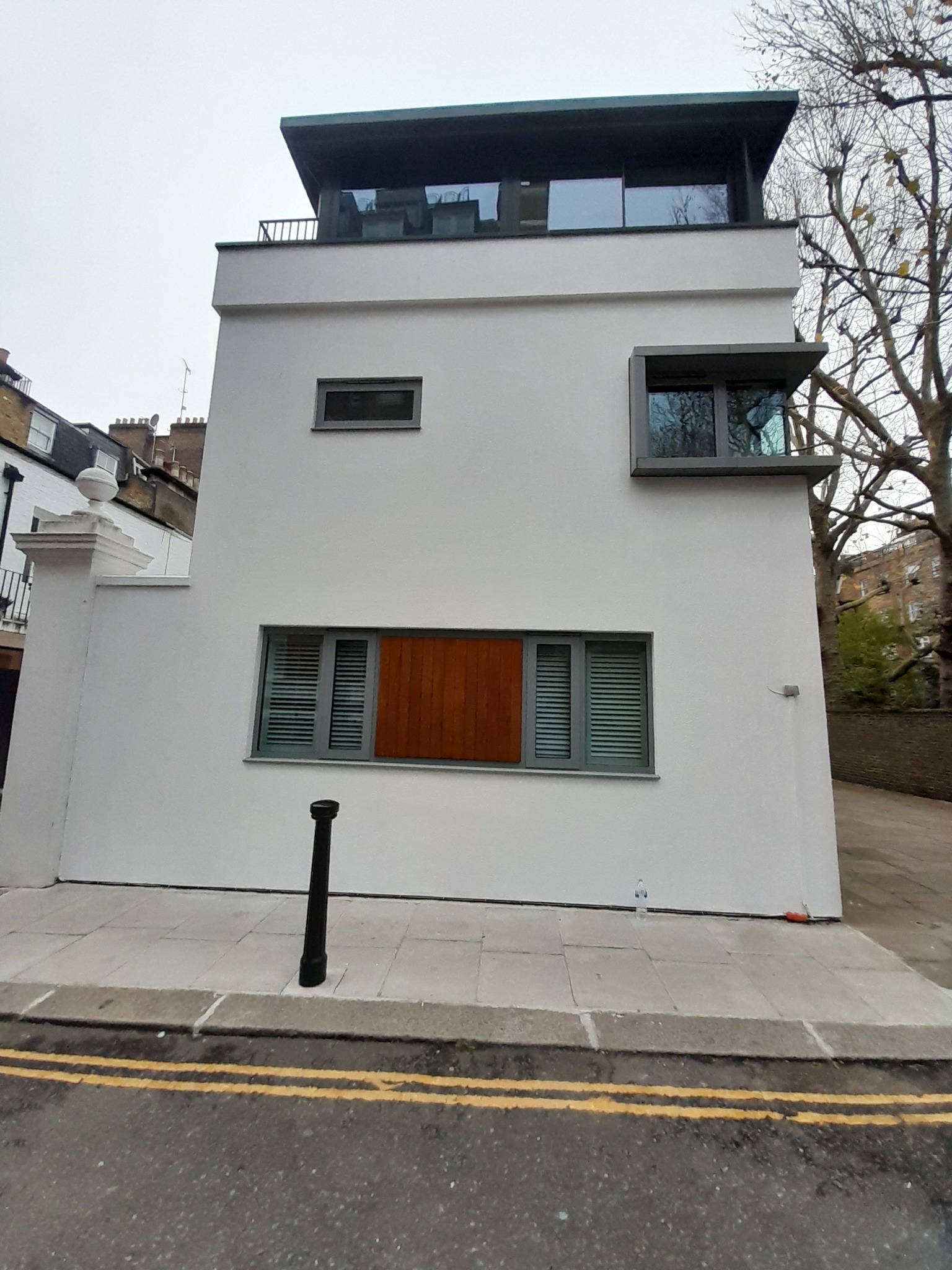 Nowoczesny, trzypiętrowy budynek pomalowany na biało z szarymi ramami okiennymi i drewnianym panelem zasłaniającym część okna, widoczny z poziomu ulicy z żółtymi liniami.