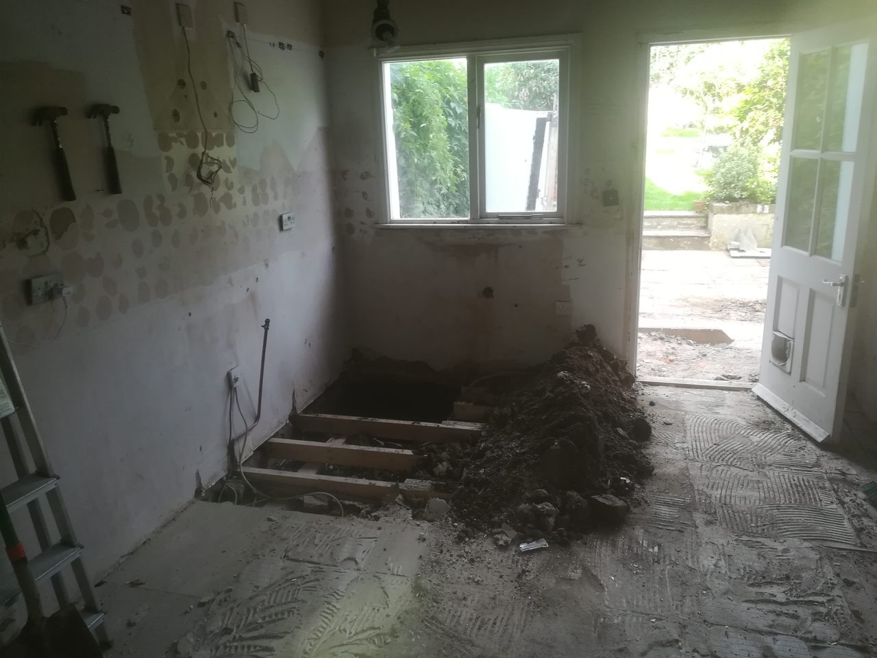Wnętrze w trakcie remontu: ściany z usuniętą farbą, odsłonięta podłoga z drewnianymi belkami, sterta ziemi przy drzwiach wejściowych, drabina oparta o ścianę, dwa młotki wiszące na ścianie.