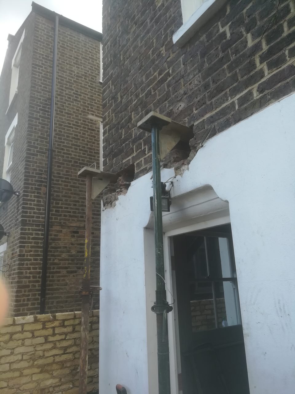 Uszkodzona ściana budynku podparta stemplami budowlanymi, widoczne braki w murze i tynku wokół ramy okiennej.