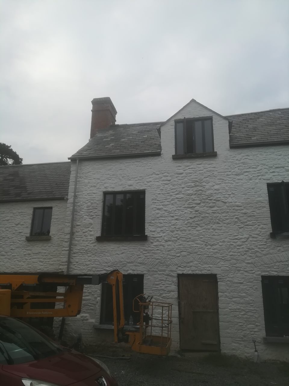 Biała, kamienna elewacja budynku po malowaniu, widoczny podnośnik koszowy i fragment samochodu, pochmurne niebo.