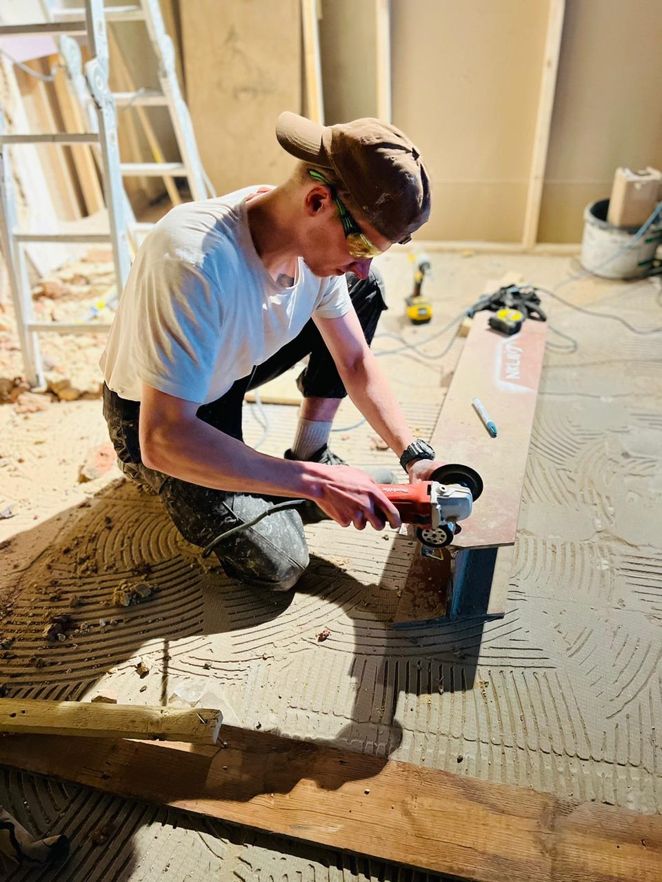 Mężczyzna w okularach ochronnych szlifuje płytkę szlifierką kątową na podłodze w trakcie remontu, widoczne narzędzia i resztki materiałów budowlanych.