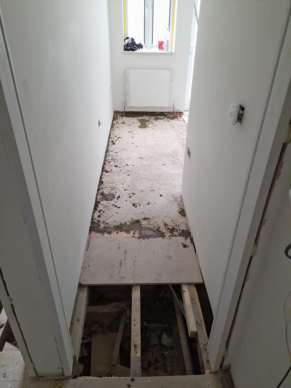 Wnętrze w trakcie remontu: zerwana podłoga, odsłonięte legary, przygotowane ściany do malowania, okno z grzejnikiem w tle.