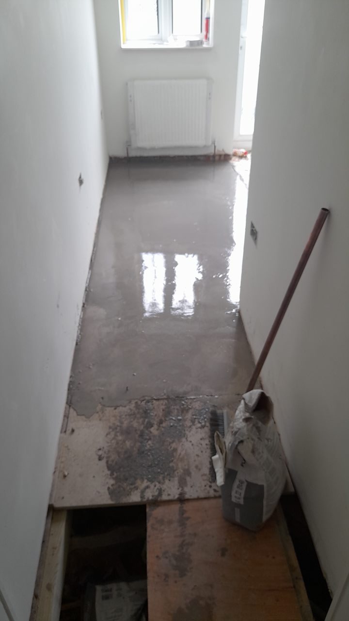 Wylewka samopoziomująca w korytarzu, widoczny worek z zaprawą, szczotka do rozprowadzania, oraz otwór w podłodze.