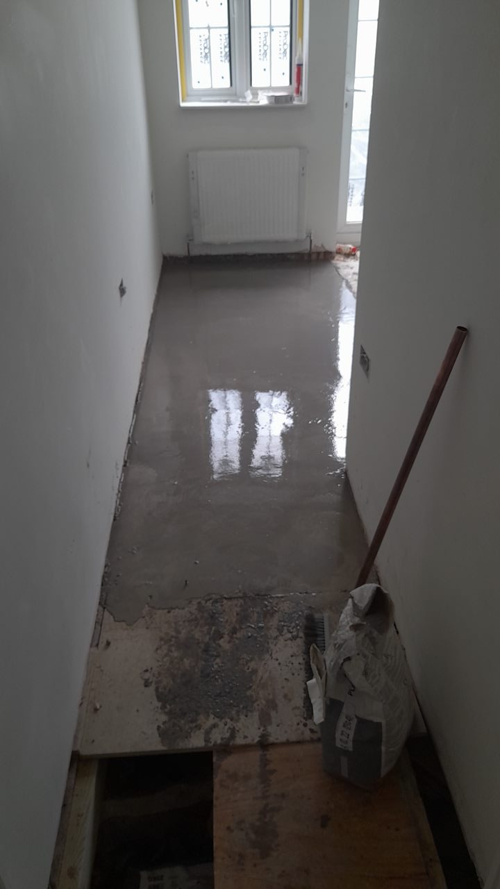 Świeżo wylana, mokra wylewka samopoziomująca w wąskim pomieszczeniu z grzejnikiem pod oknem, widoczny worek z zaprawą i rurka oparta o ścianę.