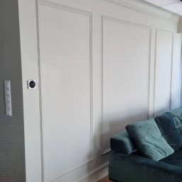 Aleksander Gutkowski ALLEL - Eleganckie tapetowanie w stylu angielskim z delikatną teksturą, listwy dekoracyjne, parkiet w jodełkę i fragment zielonej sofy.