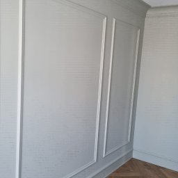 Aleksander Gutkowski ALLEL - Ściana wykończona w jasnoszarym kolorze z dekoracyjnymi panelami i wzorzystą strukturą, uzupełniona parkietem ułożonym w jodełkę.