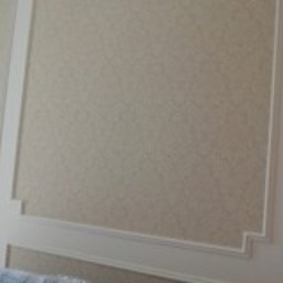 Aleksander Gutkowski ALLEL - Ściana wytapetowana w jasnym, beżowym odcieniu z delikatnym wzorem, ozdobiona białymi, dekoracyjnymi listwami tworzącymi prostokątne panele.