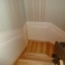 Aleksander Gutkowski ALLEL - Klatka schodowa z drewnianymi schodami i ścianami wykończonymi panelami oraz tapetą w pionowe pasy w górnej części.