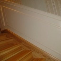 Aleksander Gutkowski ALLEL - Fragment klatki schodowej z drewnianymi schodami i pomalowaną na biało boazerią ścienną z delikatnym frezowaniem.