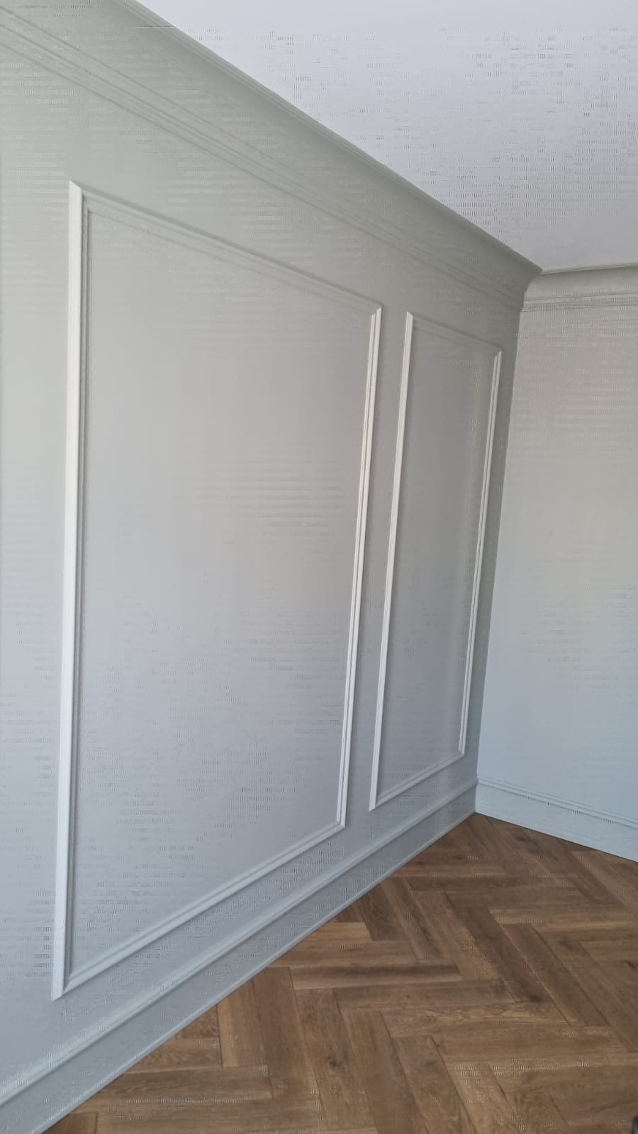 Ściana wykończona w jasnoszarym kolorze z dekoracyjnymi panelami i wzorzystą strukturą, uzupełniona parkietem ułożonym w jodełkę.