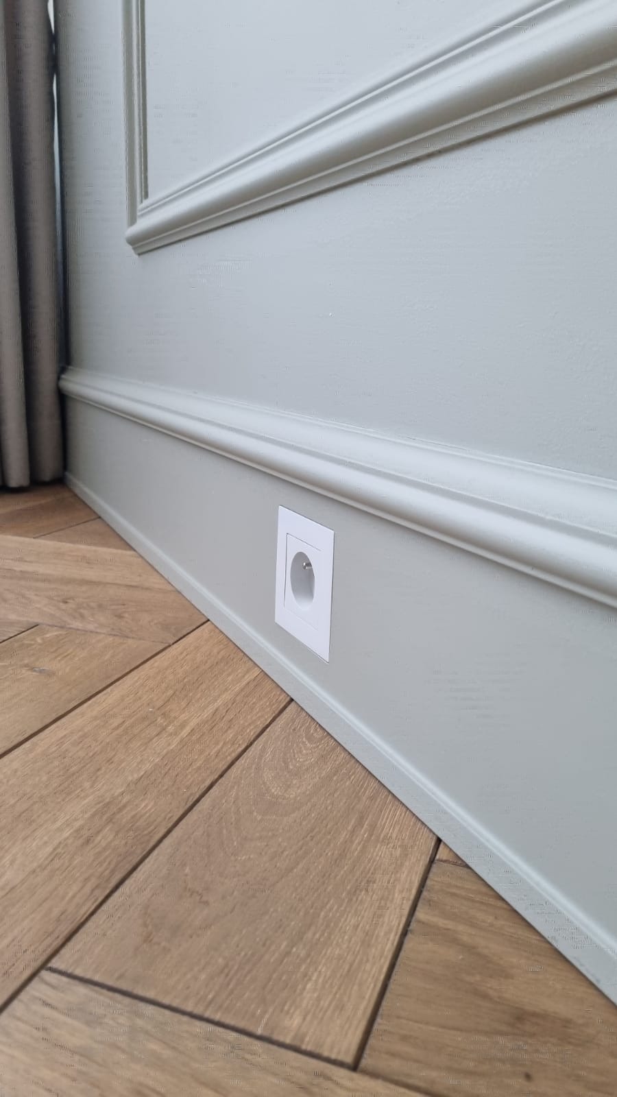 Ściana w kolorze gołębiej szarości z dekoracyjną listwą przypodłogową i gniazdkiem elektrycznym, parkiet ułożony w jodełkę.