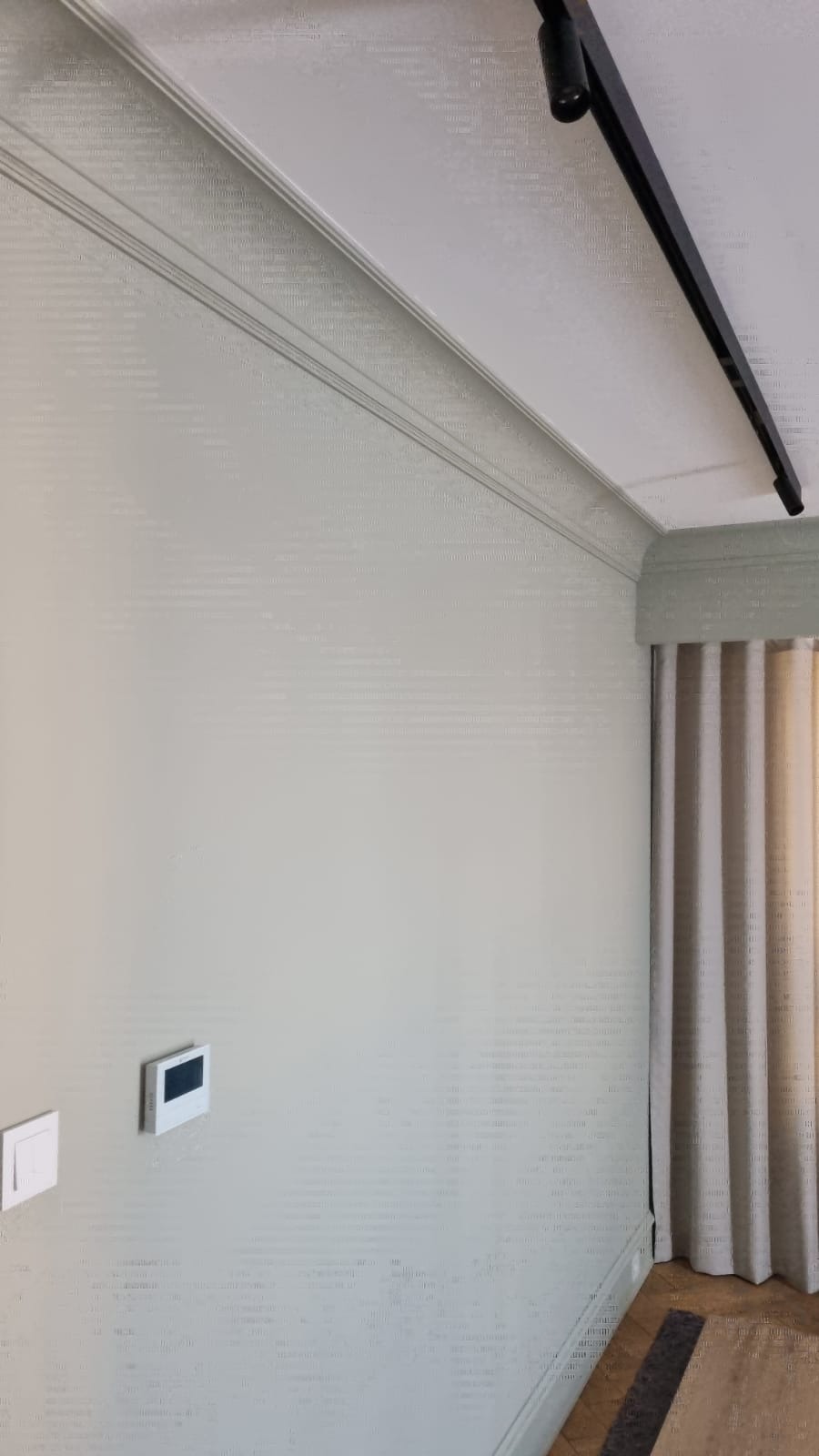 Jasnoszara ściana z trzema listwami dekoracyjnymi pod sufitem, czarnym reflektorem szynowym i zasłoniętym oknem z beżowymi zasłonami