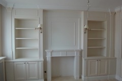 Białe, wbudowane regały z szafkami i półkami po bokach atrapy kominka w jasnym pomieszczeniu.