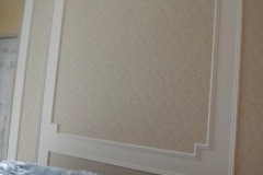 Ściana wytapetowana w jasnym, beżowym odcieniu z delikatnym wzorem, ozdobiona białymi, dekoracyjnymi listwami tworzącymi prostokątne panele.