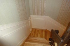 Klatka schodowa z drewnianymi schodami i ścianami wykończonymi panelami oraz tapetą w pionowe pasy w górnej części.