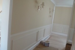 Korytarz z kremowymi ścianami, białymi panelami dekoracyjnymi i drewnianą podłogą. Widoczne kinkiety w stylu retro i puszki z farbą w rogu.