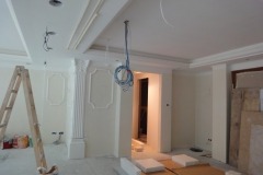 Wnętrze w trakcie wykańczania, widoczne sztukaterie na suficie i ścianach, drabina oparta o ścianę, przewody zwisające z sufitu, przygotowane płyty styropianowe na podłodze.