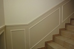 Klatka schodowa z kremowymi ścianami ozdobionymi białymi panelami dekoracyjnymi i brązowymi płytkami na schodach.