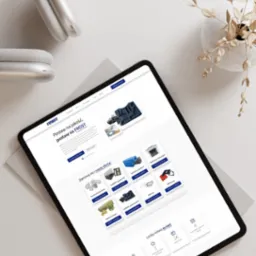 Tablet wyświetla stronę internetową firmy Frost z ofertą wyrobów styropianowych i piankowych, słuchawki bezprzewodowe i wazonik z suszem na białym stole w neutralnym oświetleniu.