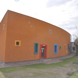 MIKOSTUDIO arch. Krzysztof Pocałujko - Pomarańczowy budynek o zaokrąglonym kształcie z kolorowymi oknami i czerwonymi drzwiami wejściowymi, widziany z zewnątrz w słoneczny dzień.