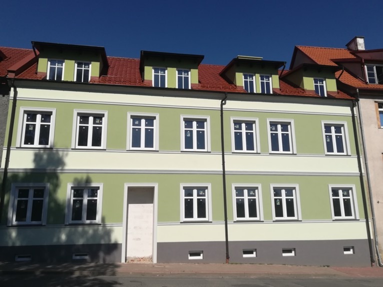 Odnowiona elewacja budynku wielorodzinnego z nowymi oknami, pomalowana na jasnozielony kolor z kremowymi akcentami, widoczne trzy okna dachowe z ciemnymi obramowaniami.
