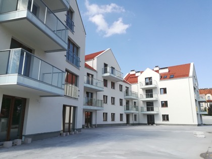 Nowoczesny, trzypiętrowy budynek apartamentowy z białymi ścianami, czerwonym dachem i szklanymi balkonami, sfotografowany w słoneczny dzień z niskiej perspektywy.